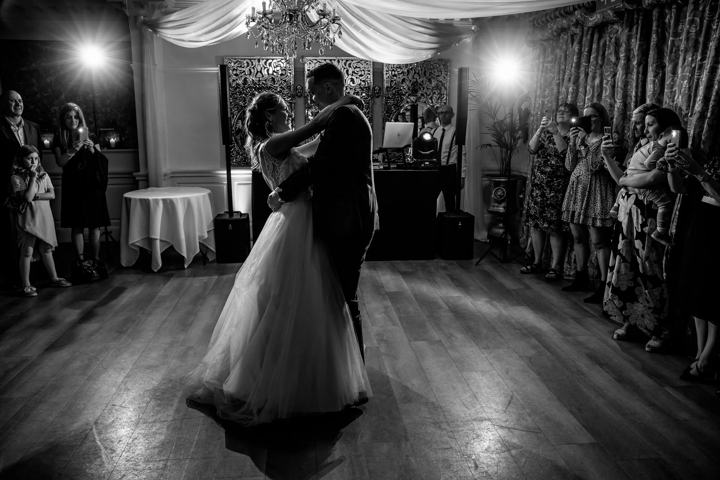 A bride and groom are dancing in the center of a wedding reception, surrounded by guests taking photos and watching. The scene is indoors with draped ceiling decor, a chandelier, and curtains. The lighting creates a silhouette effect around the coupl