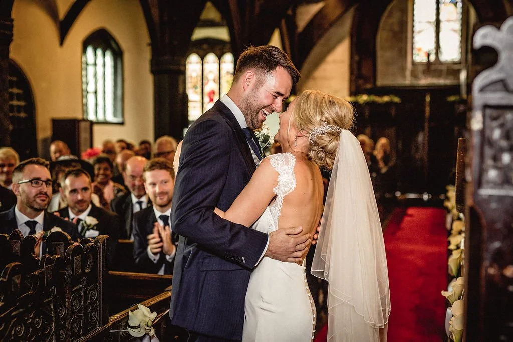 A bride and groom share a dance and smile at their wedding reception in a church with guests clapping and smiling in the background.