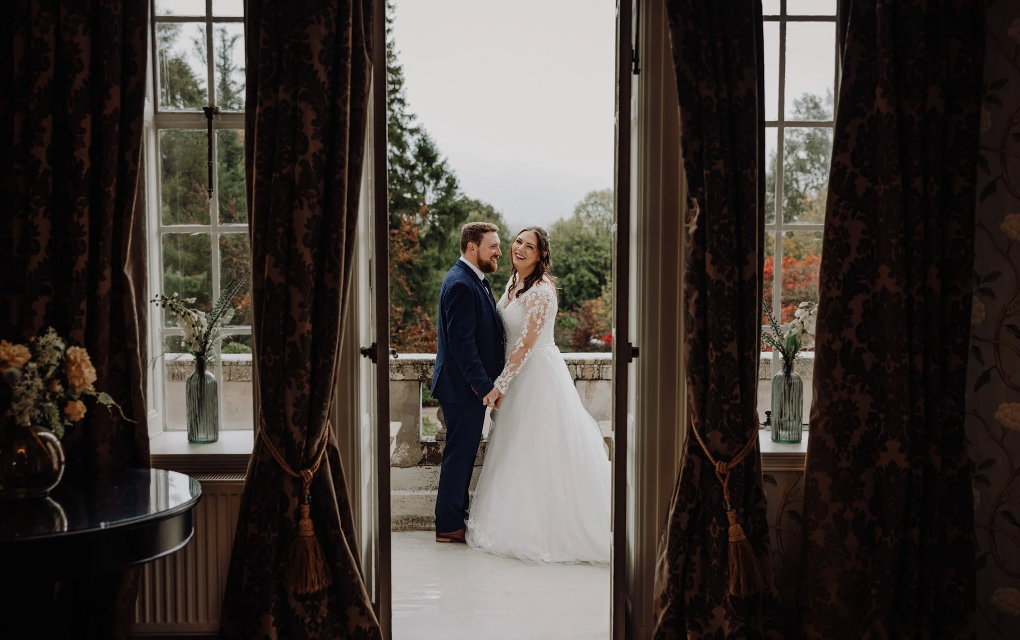 A bride and groom smiling and holding hands on a balcony, seen through open curtains, with a view of trees and sky outside.
