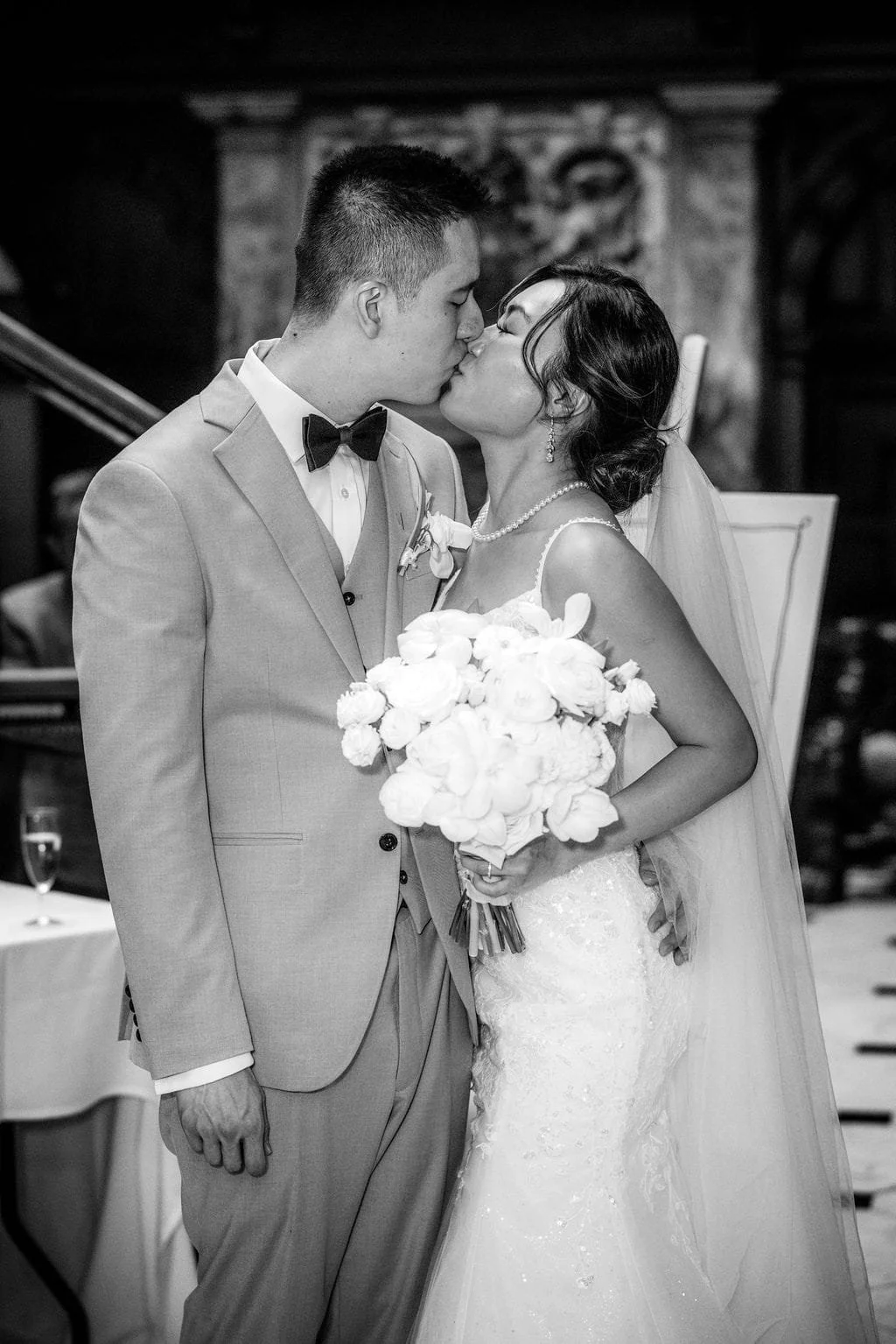 A black and white photo of a newlywed couple sharing a kiss during their wedding ceremony. The groom is in a light-colored suit with a bow tie, and the bride is holding a bouquet of flowers, wearing a wedding dress and veil.