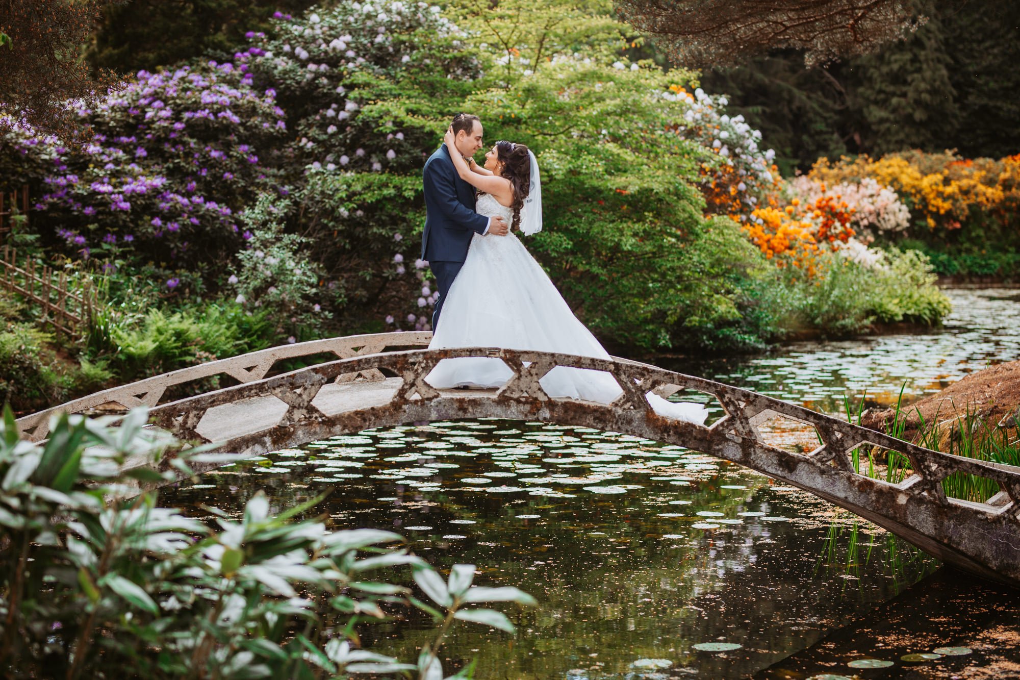 A bride and groom standing on a small stone bridge over a pond, surrounded by colorful blooming bushes and flowers.