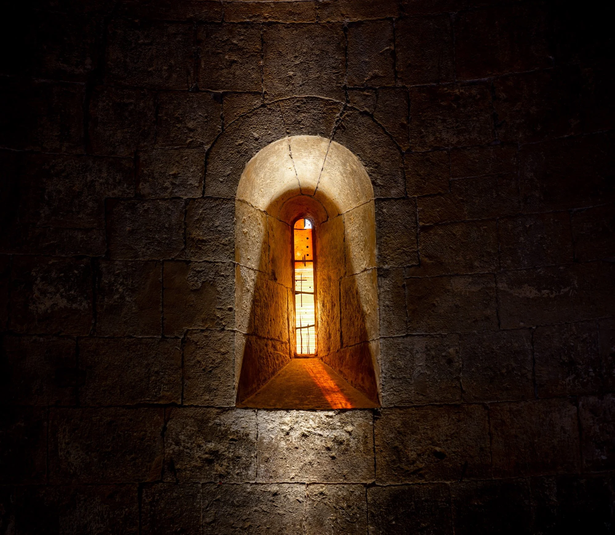 A narrow stone window in a brick wall with warm orange light shining through.