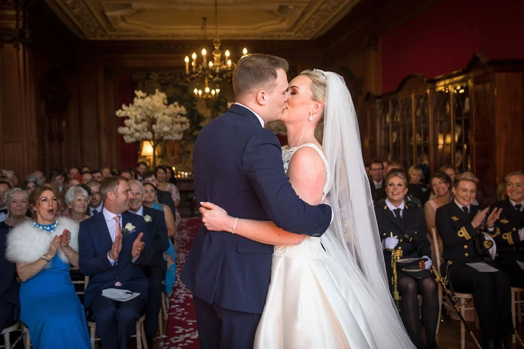 A bride and groom share a kiss during their wedding ceremony in an elegant, wood-paneled room with a chandelier and guests seated and clapping.