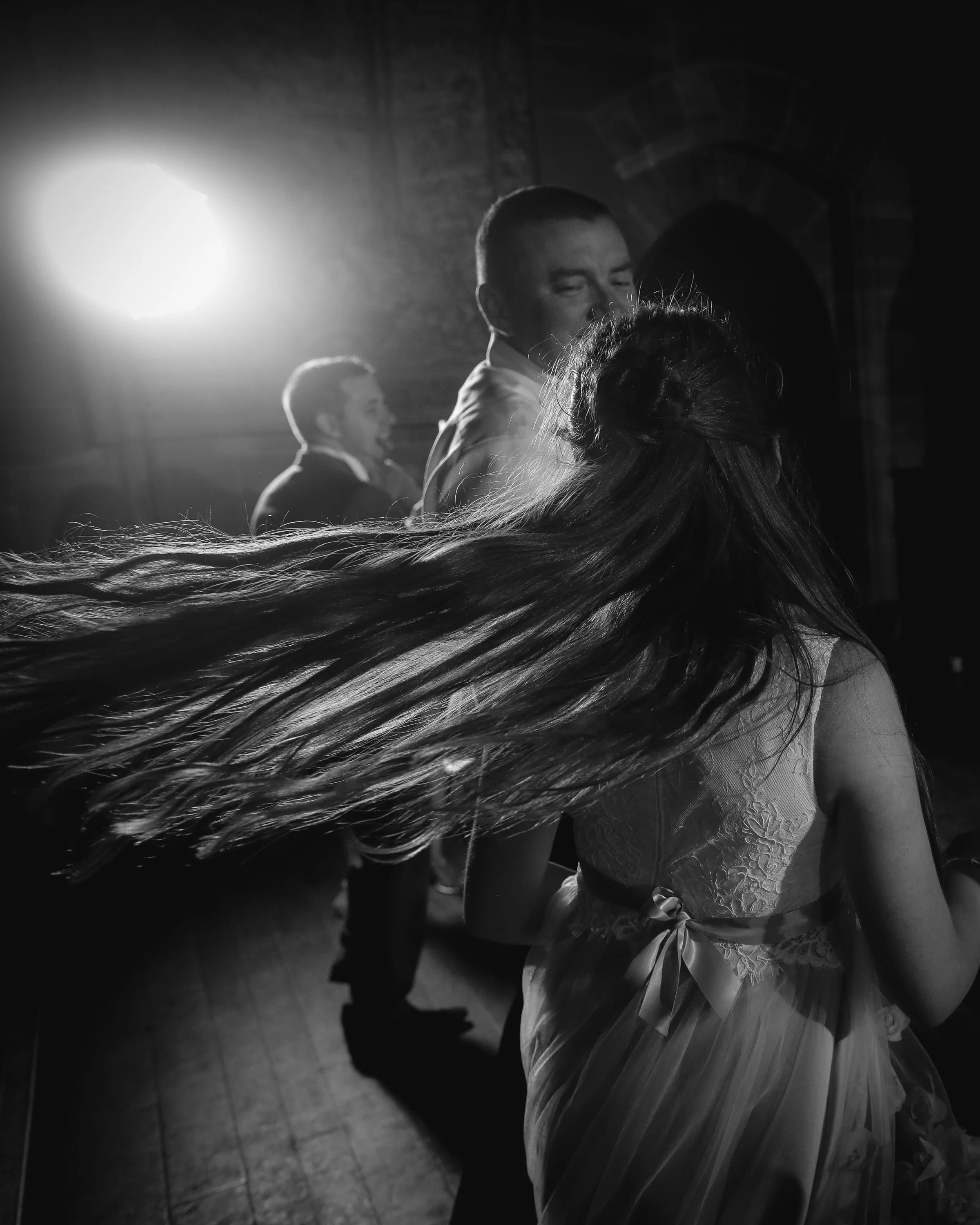 Black and white photo of people dancing indoors with a bright light in the background, featuring a woman with long hair moving in front of two men in formal attire.
