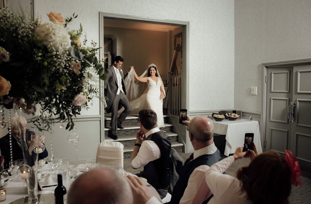 A bride and groom walking down the steps into a reception area, with guests seated at tables taking photos of them. The bride is wearing a white wedding gown and a crown, and the groom is in a gray suit. The setting is decorated for a wedding celebra