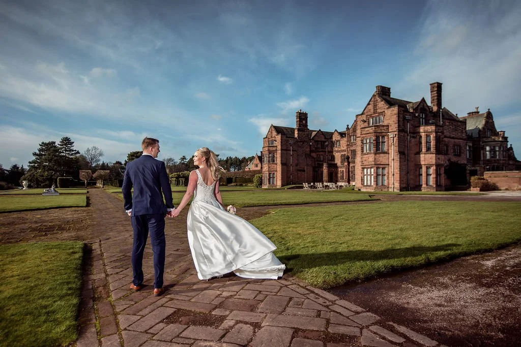 A newlywed couple walking hand in hand along a brick pathway outside a large historic mansion or castle on a partly cloudy day.