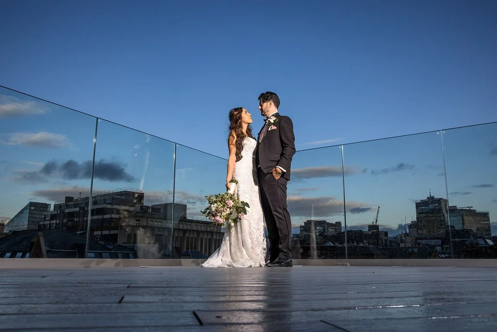 A bride and groom in wedding attire standing on a rooftop deck with the city skyline in the background, under a blue sky with a few clouds.