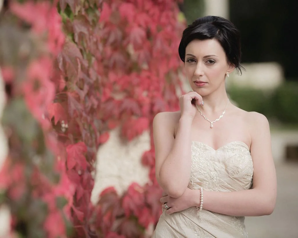 A woman in an elegant strapless dress with jewelry, standing near a wall with pink and green leaves, outdoors.