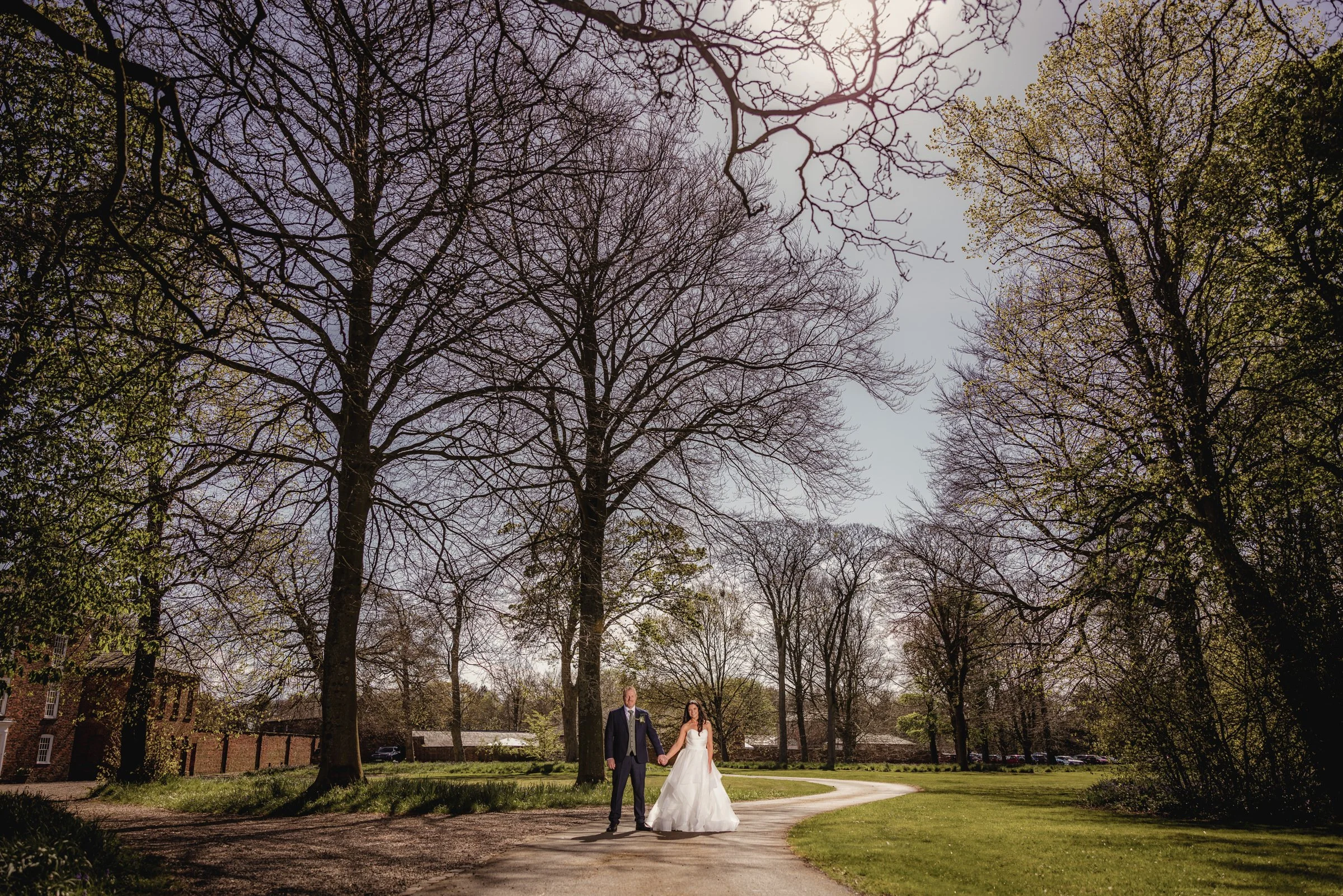 A bride and groom walking hand in hand on a winding path in a park with trees and grass on a sunny day.