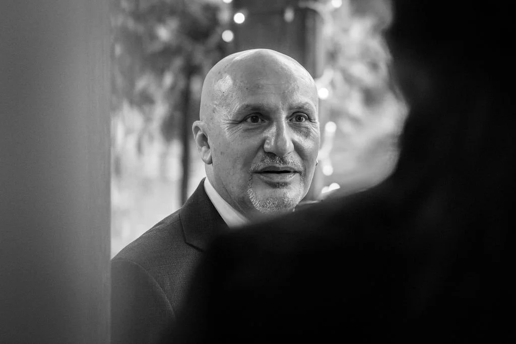 A black and white photo of a man with a bald head, dressed in a suit, looking at someone partially visible in the foreground. There are blurred lights or decorations in the background.