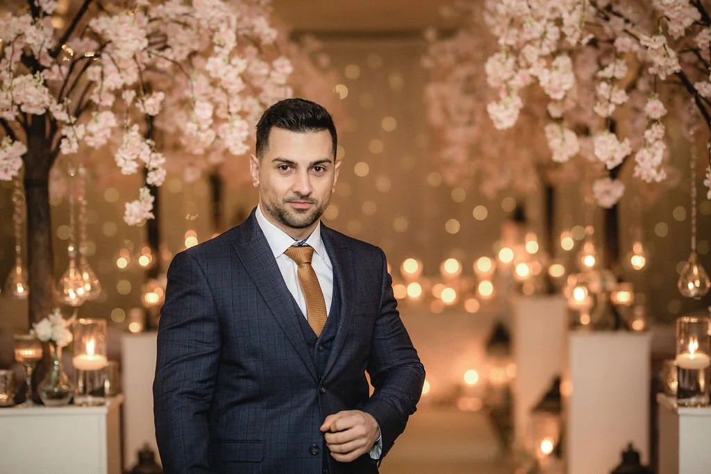 A man in a dark blue suit with a gold tie standing in a decorated indoor setting with pink cherry blossom trees and candles.