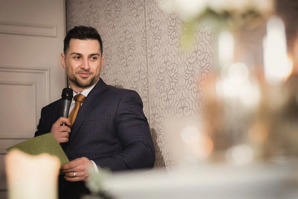 A man in a suit holding a microphone and a green paper, standing near a decorated wall, at a formal event or speech.