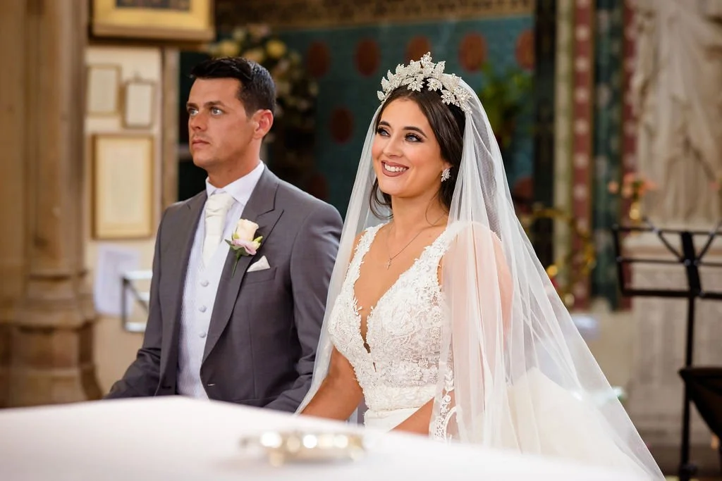 Bride and groom in wedding ceremony, bride wearing a lace wedding dress with veil and tiara, smiling; groom in a gray suit with boutonniere, looking serious, in a decorated church.