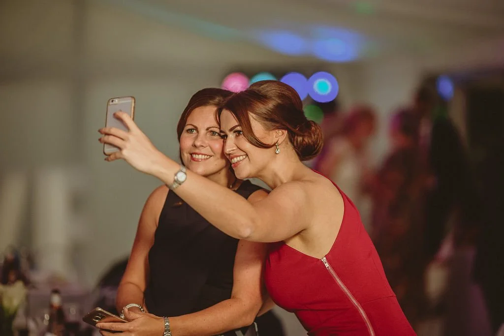 Two women taking a selfie at a social event, with one woman in a red dress and the other in a black dress, smiling and posing together.