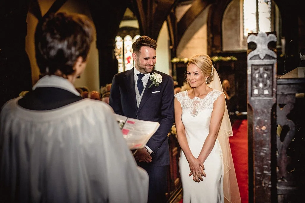 A bride and groom exchanging vows during their wedding ceremony in a church, with a priest reading from a book in front of them.