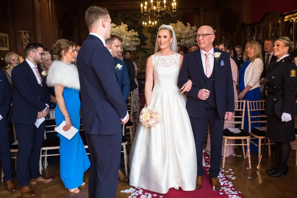 A bride in a white wedding gown with a tiara and holding a bouquet of pink flowers, walking her father down the aisle during a wedding ceremony in an elegant, wood-paneled room with floral arrangements and guests watching.