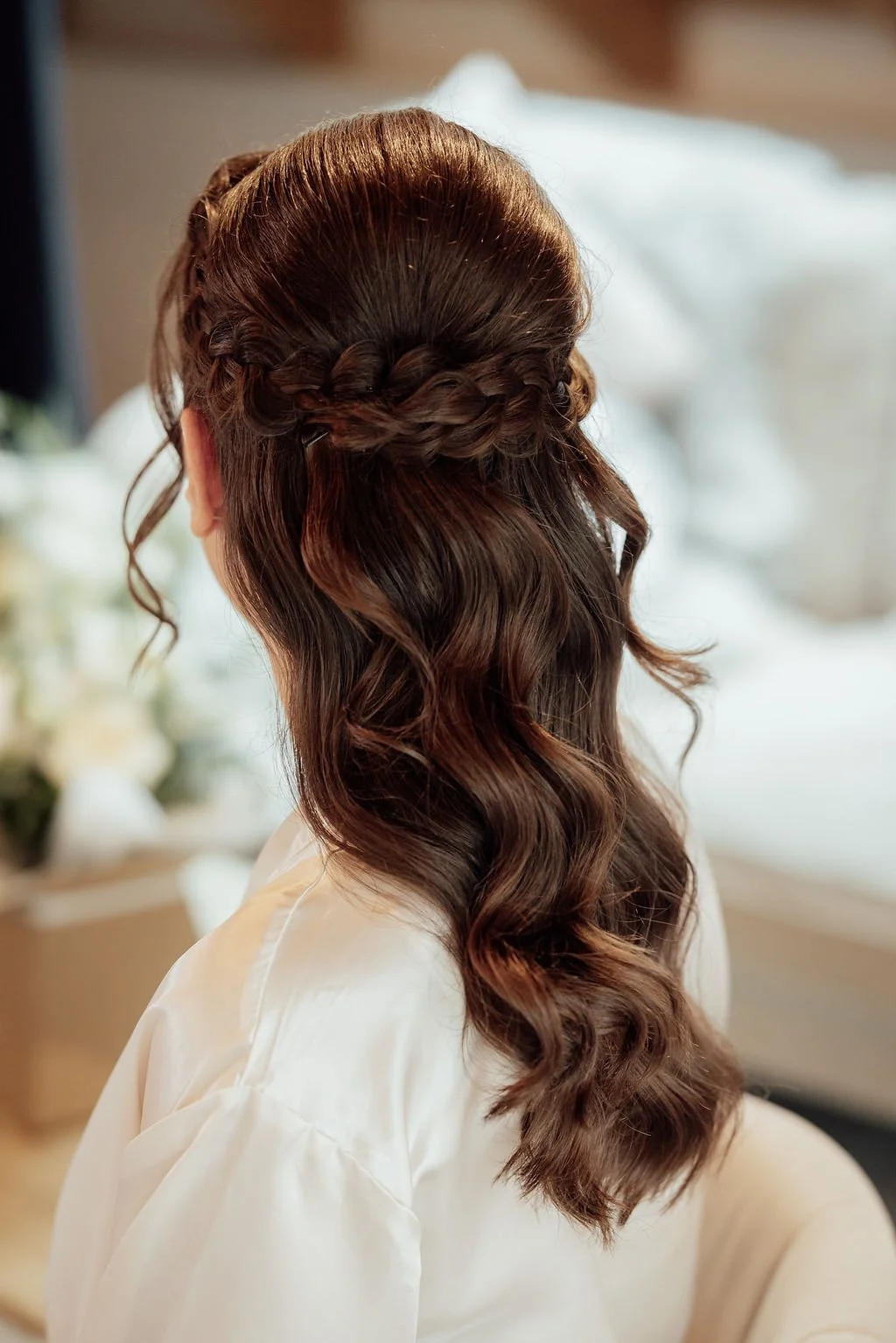 Back view of a woman with wavy brown hair styled with a braided crown and loose curls, wearing a white robe.