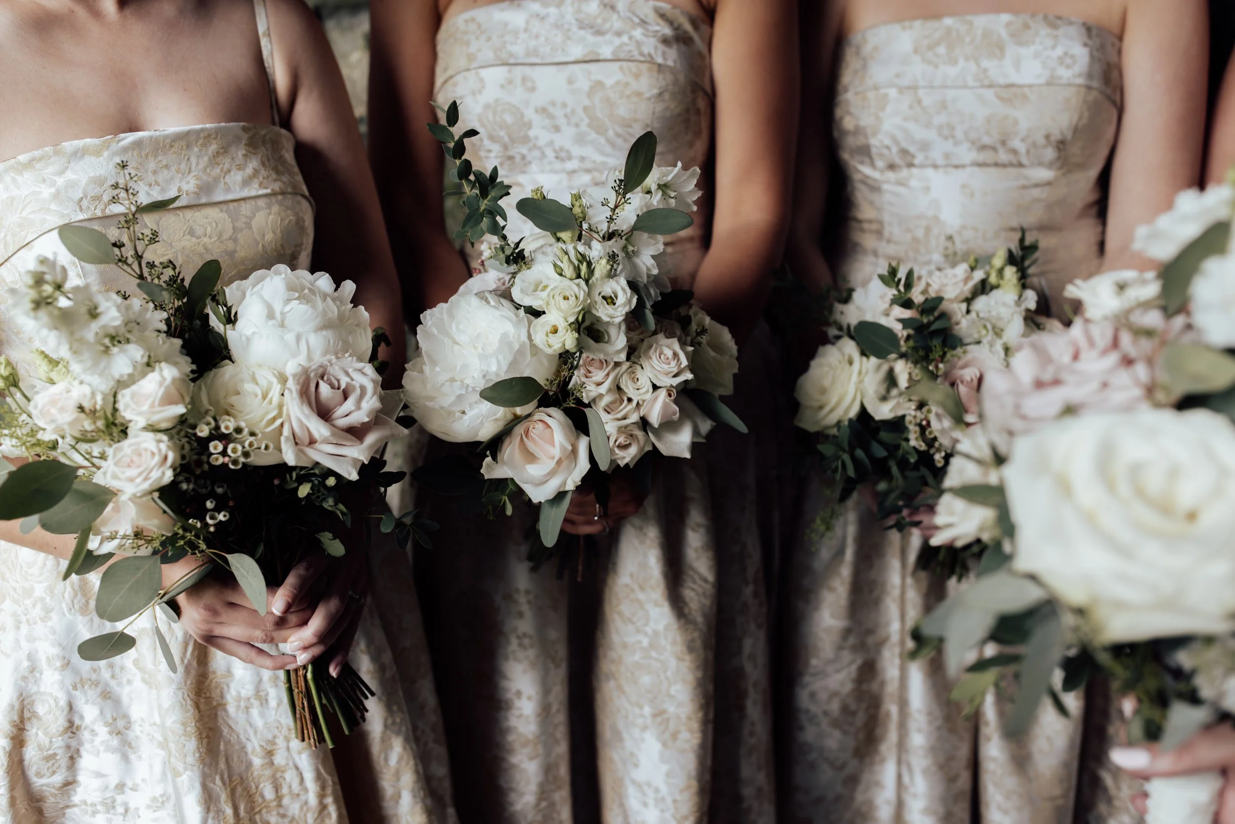 Close-up of women in lace dresses holding bouquets of white and pale pink flowers with greenery.