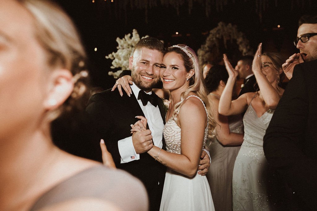A bride and groom dancing at their wedding reception, surrounded by wedding guests, with festive lighting and decorations in the background.