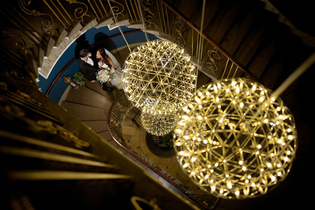 A bride and groom standing at the bottom of a spiral staircase, looking at each other, with the bride holding a bouquet of flowers, surrounded by hanging spherical chandeliers.