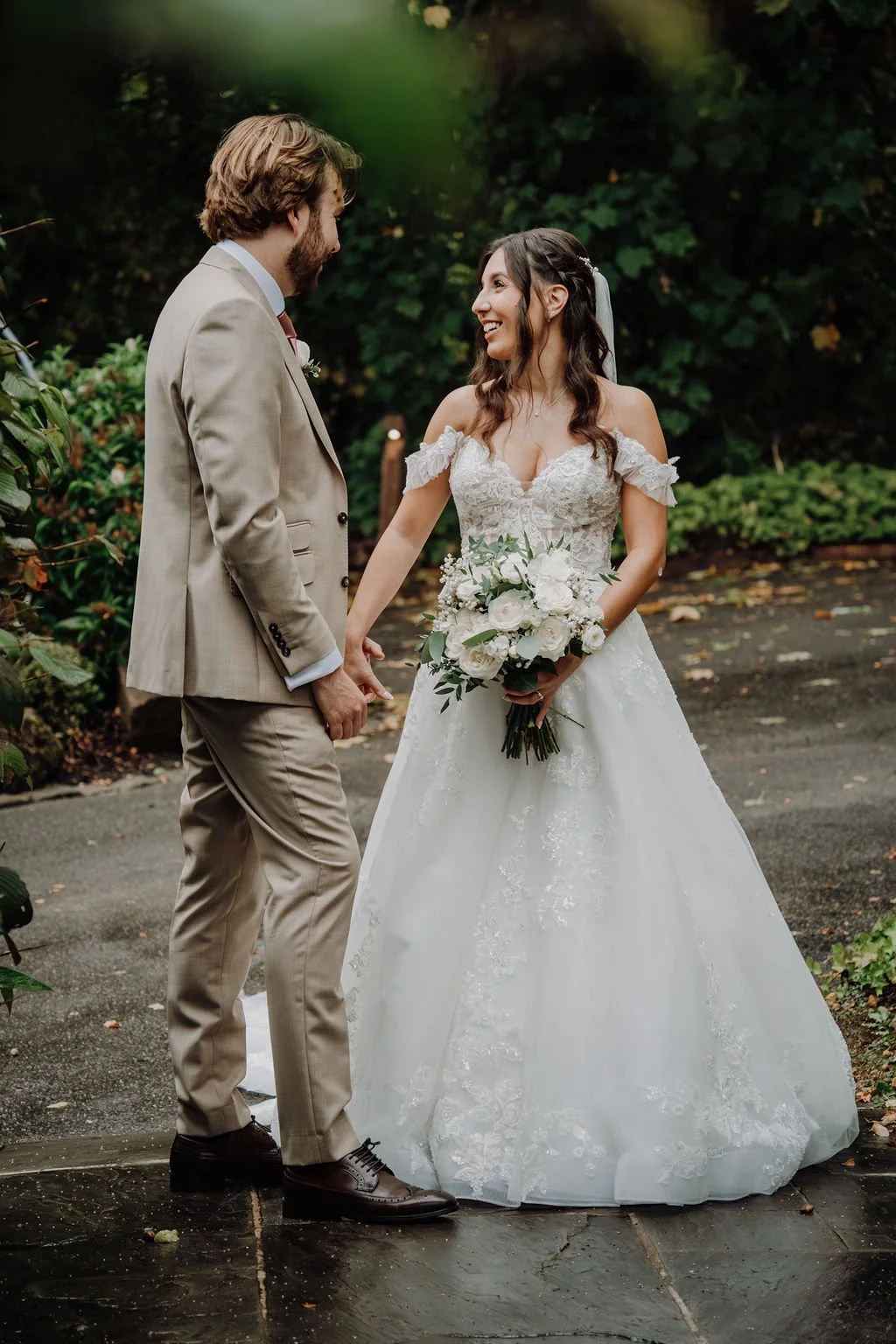 A bride and groom holding hands and smiling at each other outdoors with greenery in the background.