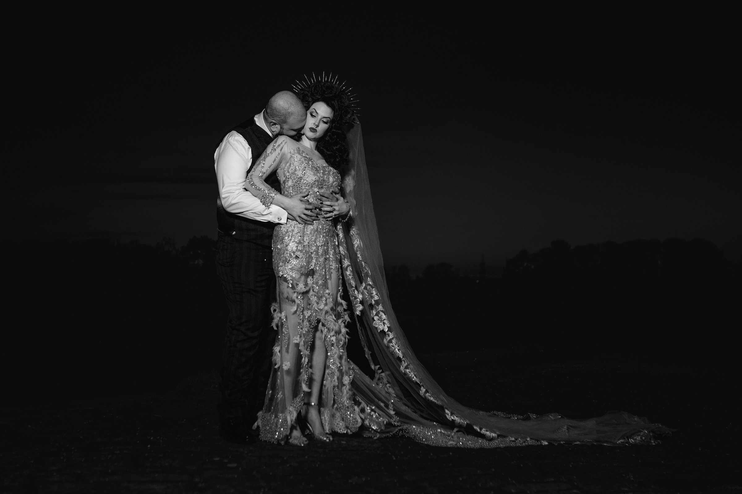 Black and white photo of a couple embracing at night, with the man kissing the woman's shoulder and the woman wearing an ornate dress and a halo-like headpiece.