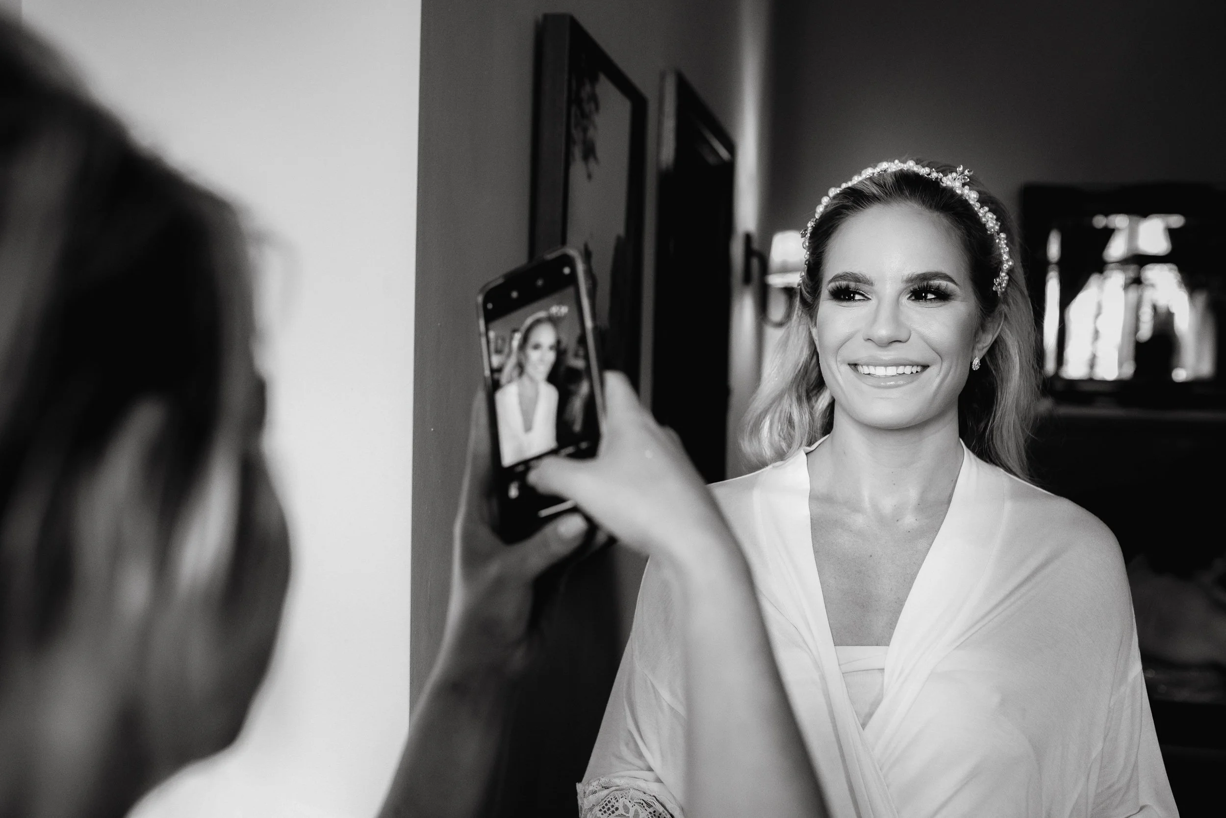 Black and white photo of a woman smiling for a photo, wearing a headband and elegant earrings, as another person captures the moment with a smartphone.