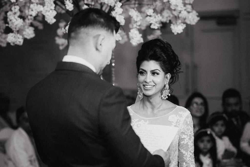 A bride and groom during their wedding vows, with the bride smiling and looking at the groom, in a decorated indoor venue with guests in the background.