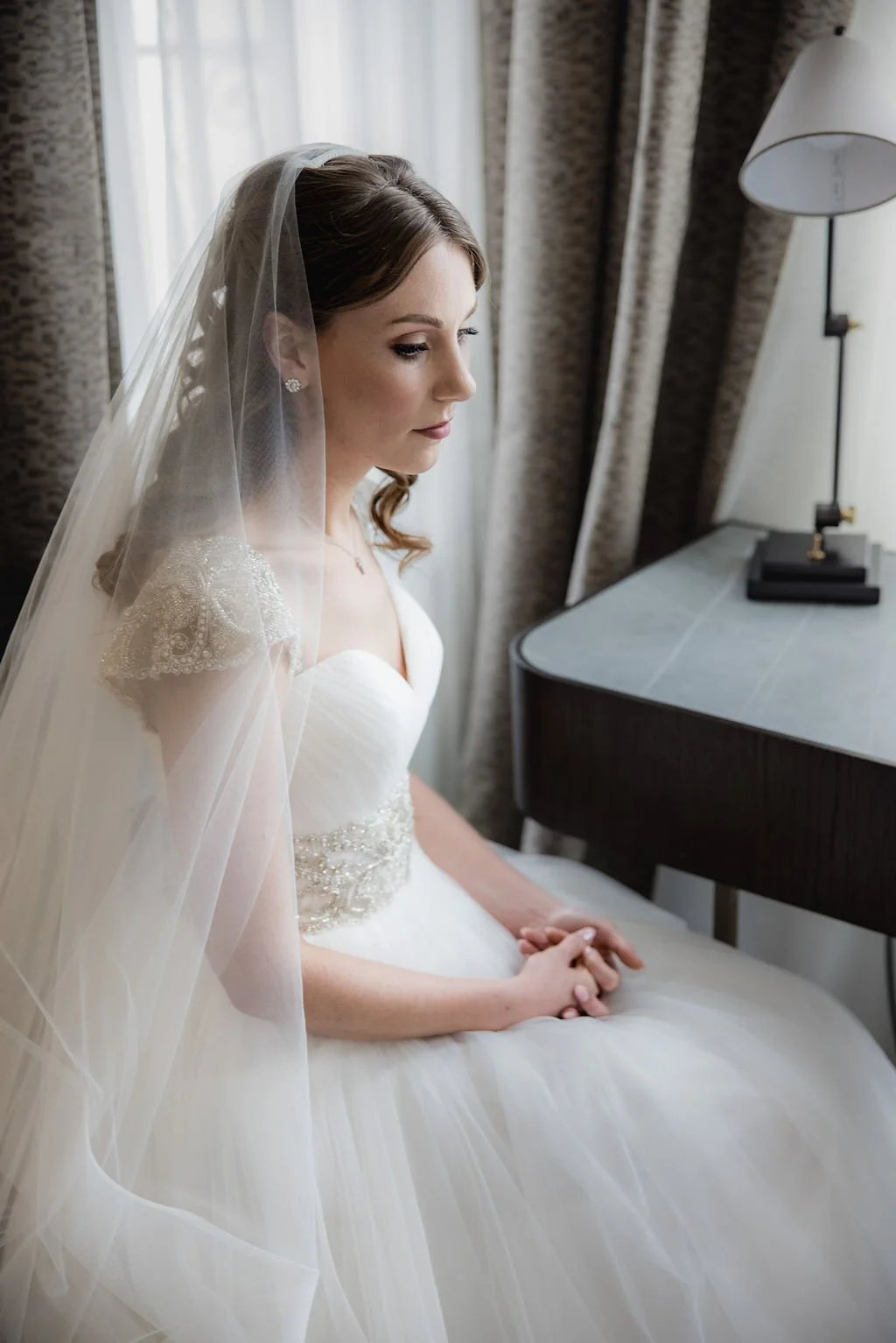 Bridal portrait of a bride with a veil and wedding gown, sitting by a window with patterned curtains and a table with a lamp.