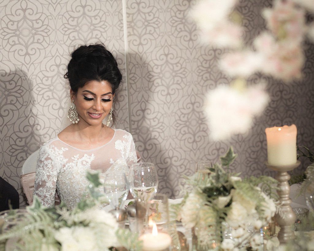 A bride in a white lace wedding dress sitting at a decorated table with floral centerpieces and candles.