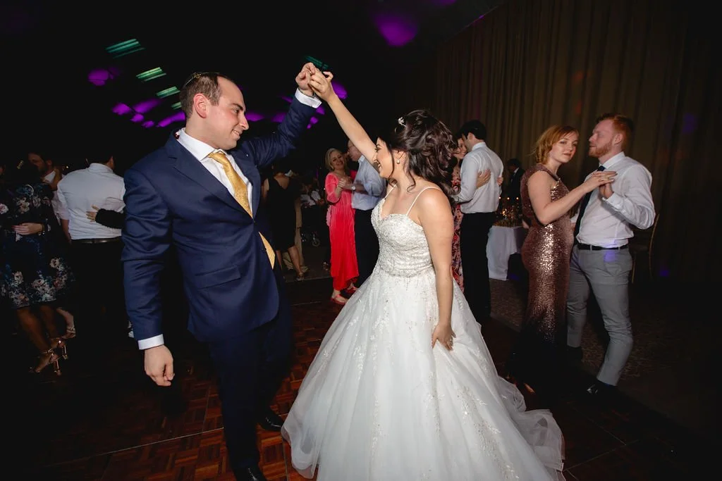 Couple dancing at a wedding reception, the groom in a navy suit with a yellow tie and the bride in a white wedding gown, with other couples dancing in the background.