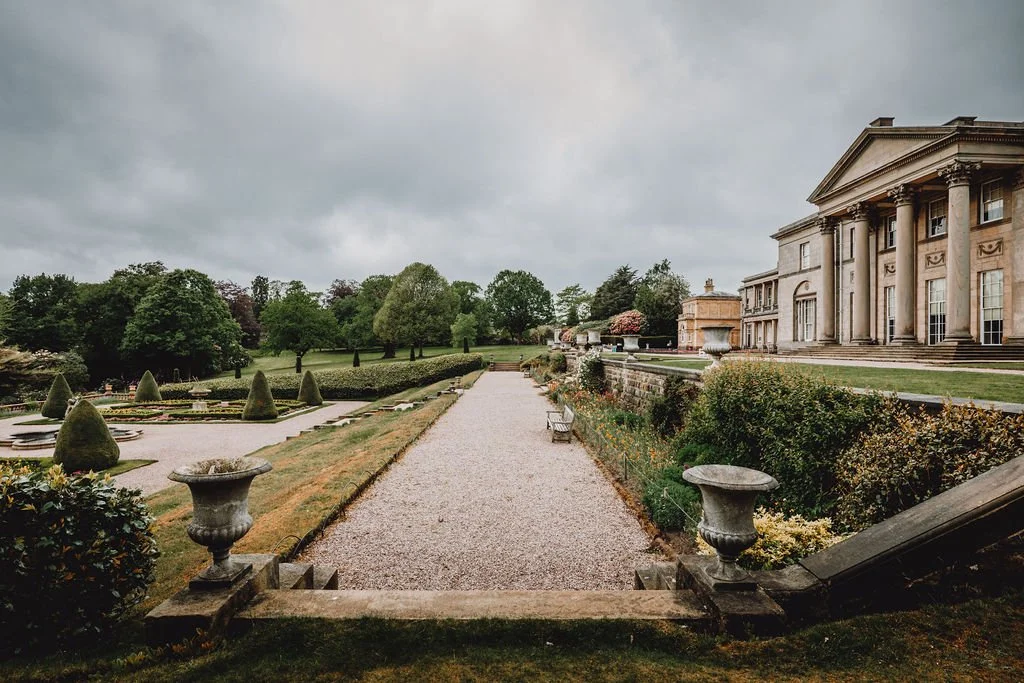 View of a grand historic mansion with columns, a gravel pathway leading to the estate, manicured gardens with topiary, and cloudy sky above.