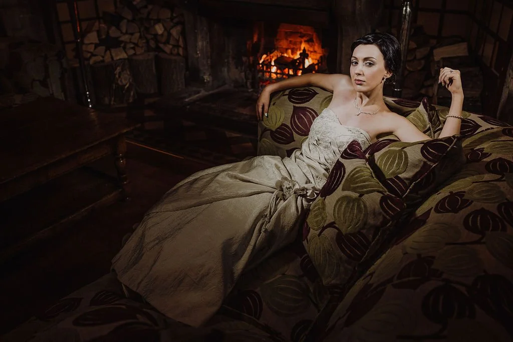 A woman with short dark hair relaxing on a patterned couch near a lit fireplace, wearing a strapless beige dress.