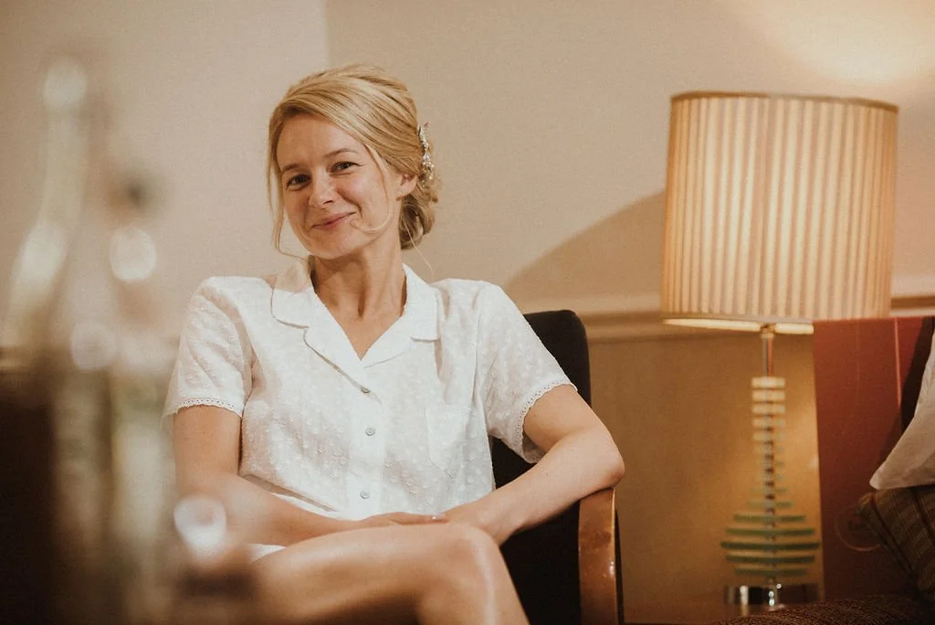 A woman with blonde hair sitting on a chair, smiling, in a room with a beige wall and a table lamp with a striped shade.