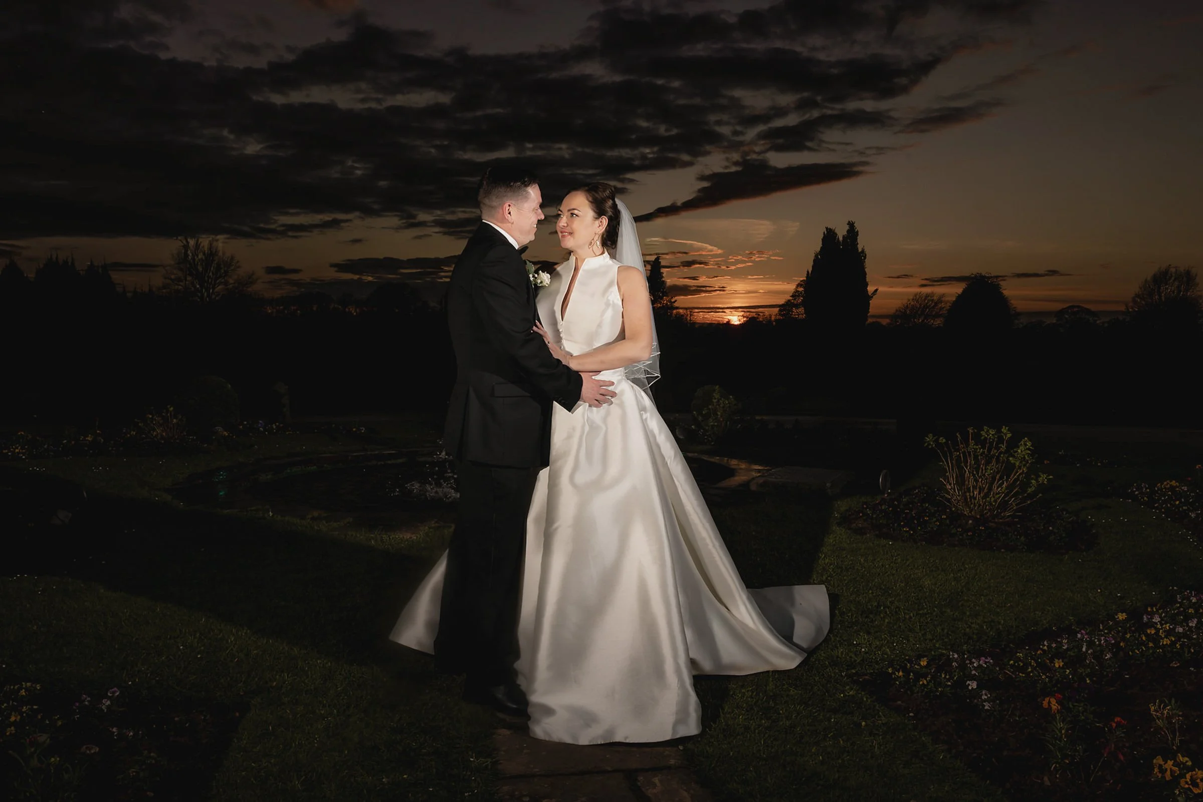 A bride and groom standing close together outdoors during sunset, with the bride wearing a white wedding gown and veil and the groom in a black tuxedo, holding hands and gazing at each other.