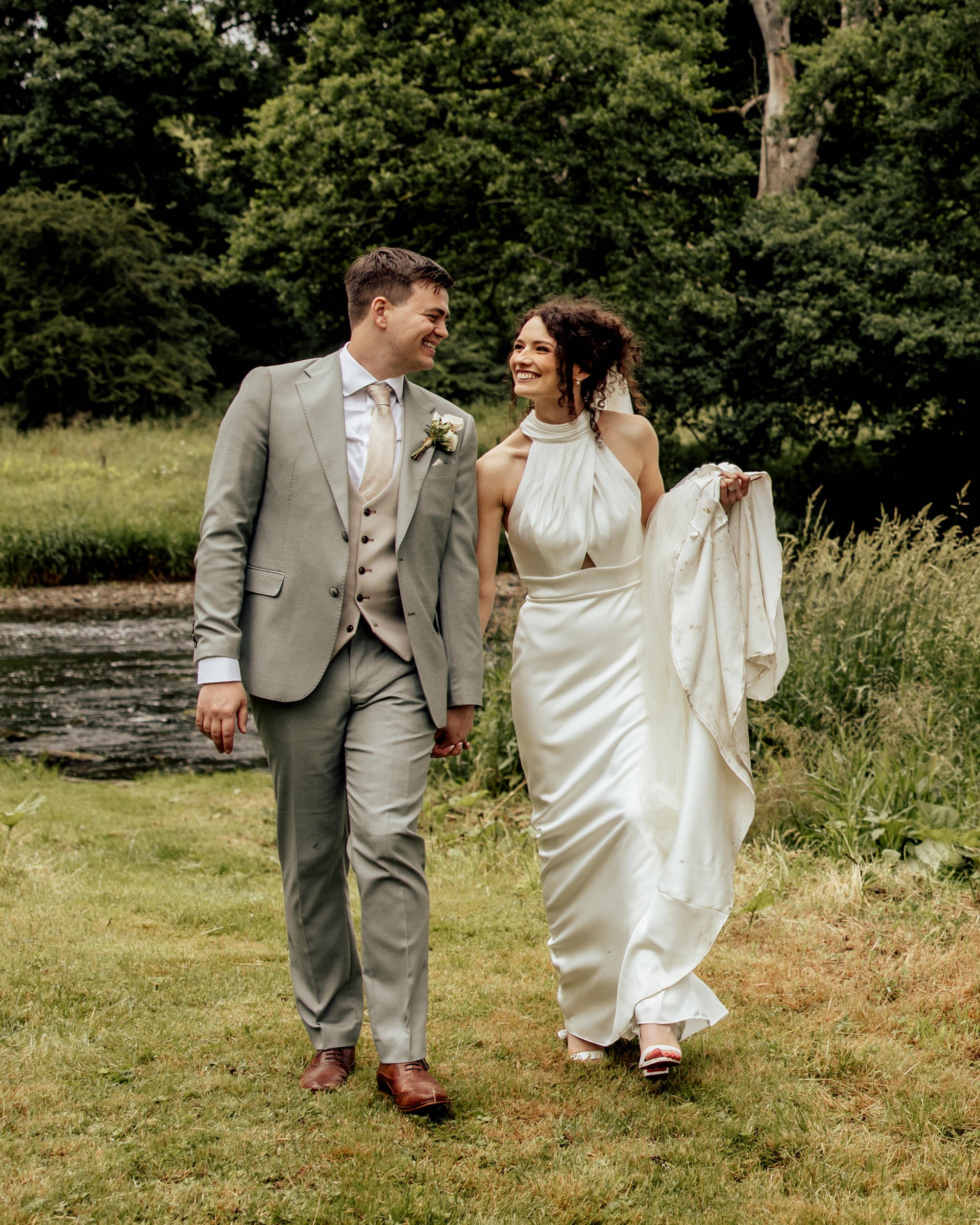 A newlywed couple walking hand in hand outdoors, smiling and looking at each other, with a lush green forest in the background.