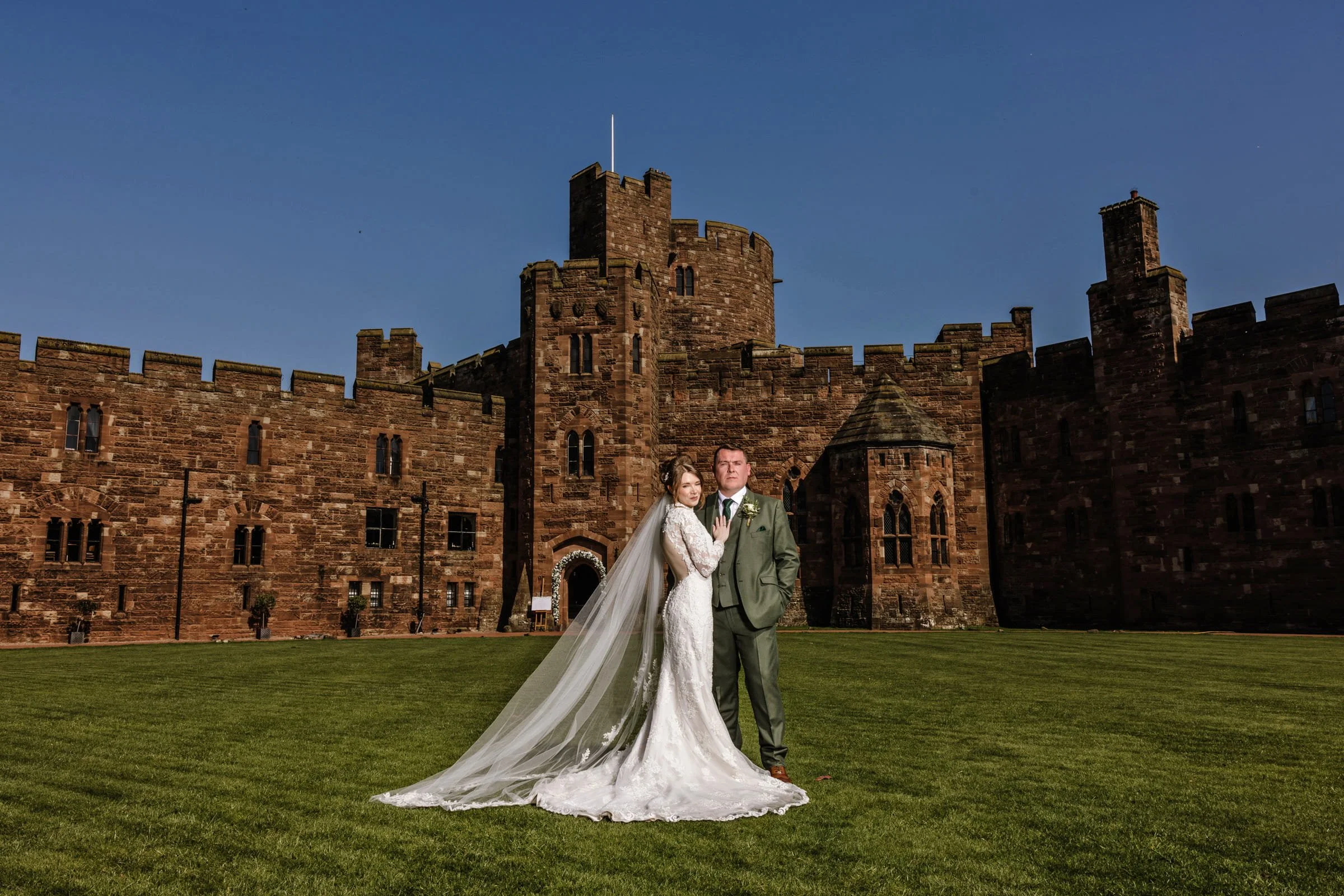 A bride and groom standing outside a castle on a sunny day. The bride is in a white wedding gown with a long train and veil, and the groom is in a green suit with a boutonniere.