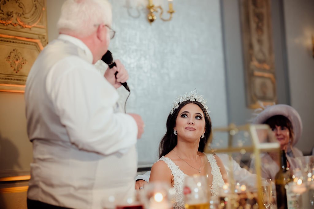 An elderly man with glasses speaking into a microphone at a wedding reception, while a bride in a lace dress and tiara listens, seated at a decorated table with another woman in a pink hat. Elegant wall decor and lighting are in the background.