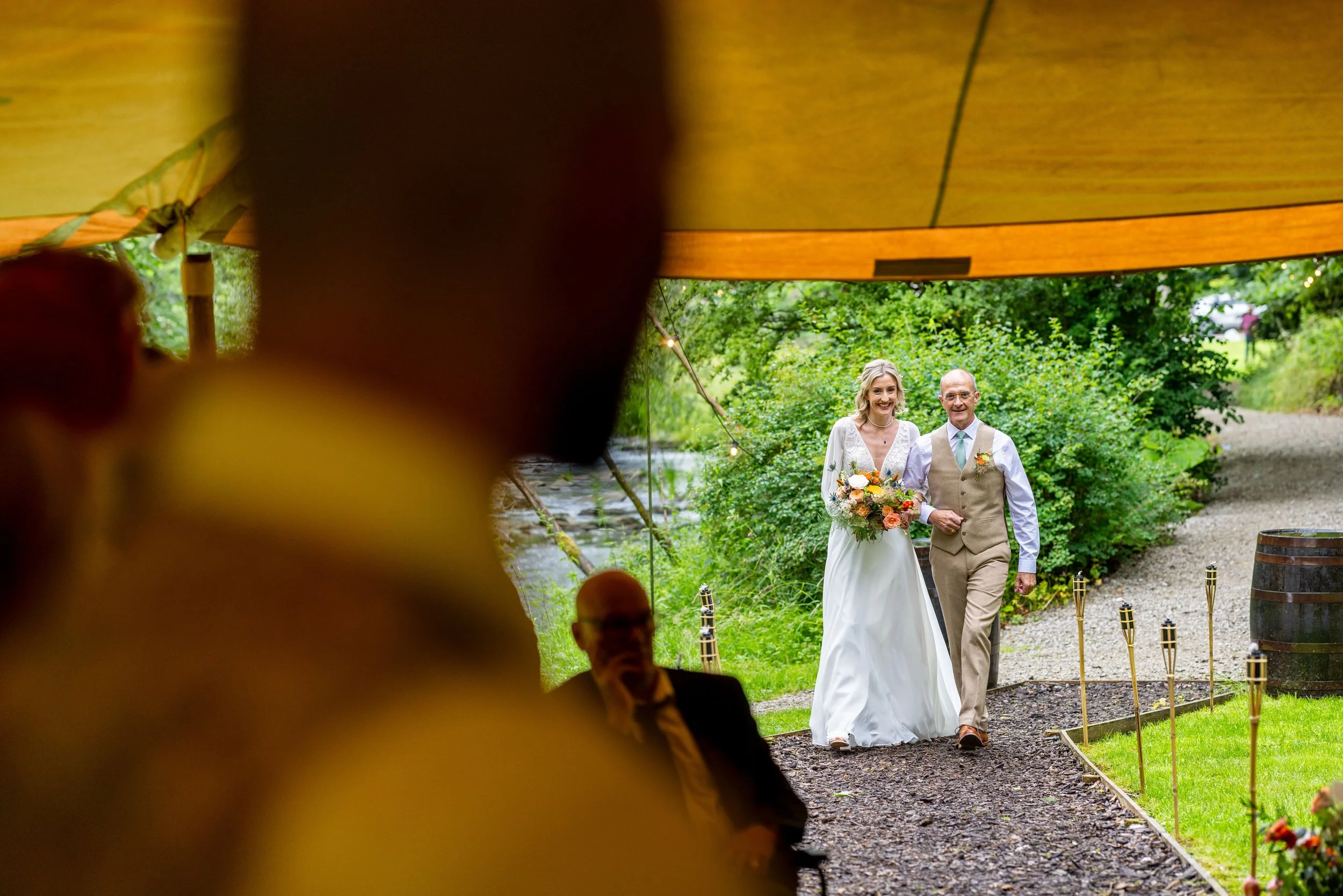 Bride walking arm-in-arm with an older man, possibly her father, under a wooden canopy at an outdoor wedding ceremony. The bride wears a white dress and holds a colorful bouquet, while the man wears a beige vest and white shirt. Green foliage and a g