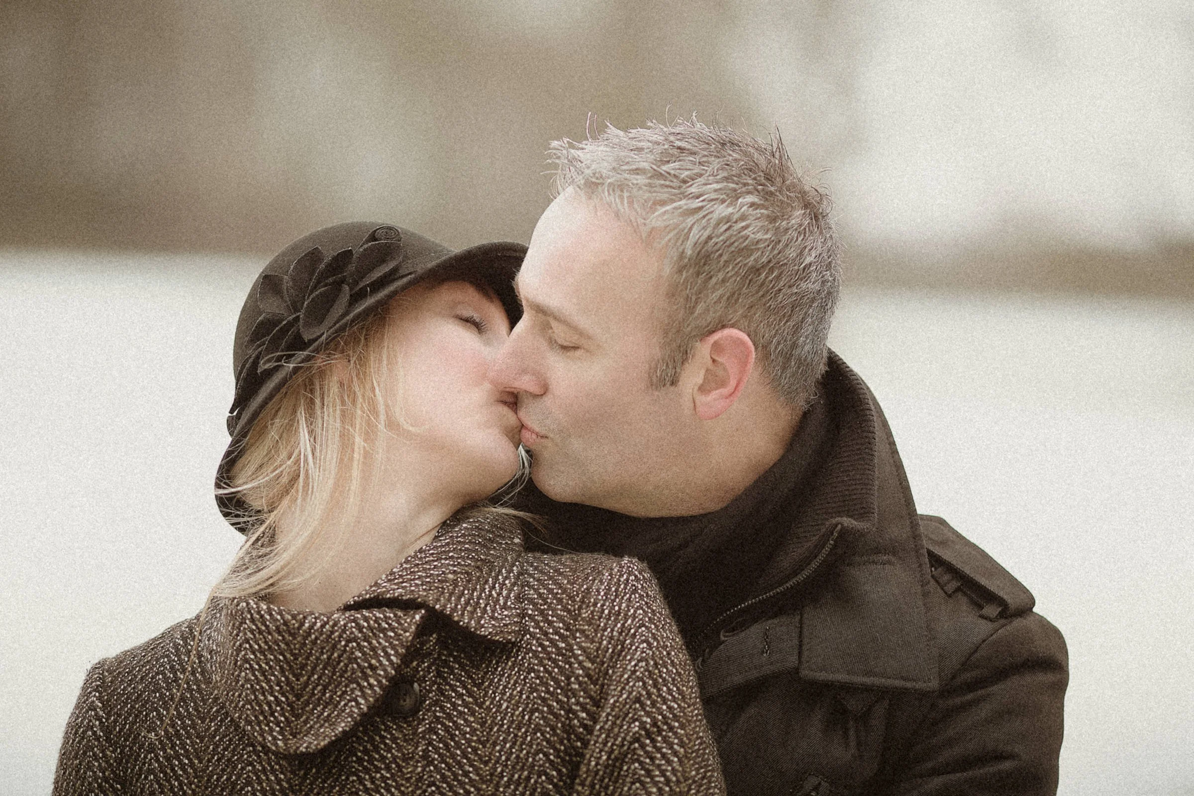 A couple kissing outdoors, with the woman wearing a black hat with decorative flowers and a brown tweed coat, and the man wearing a black jacket with a high collar.