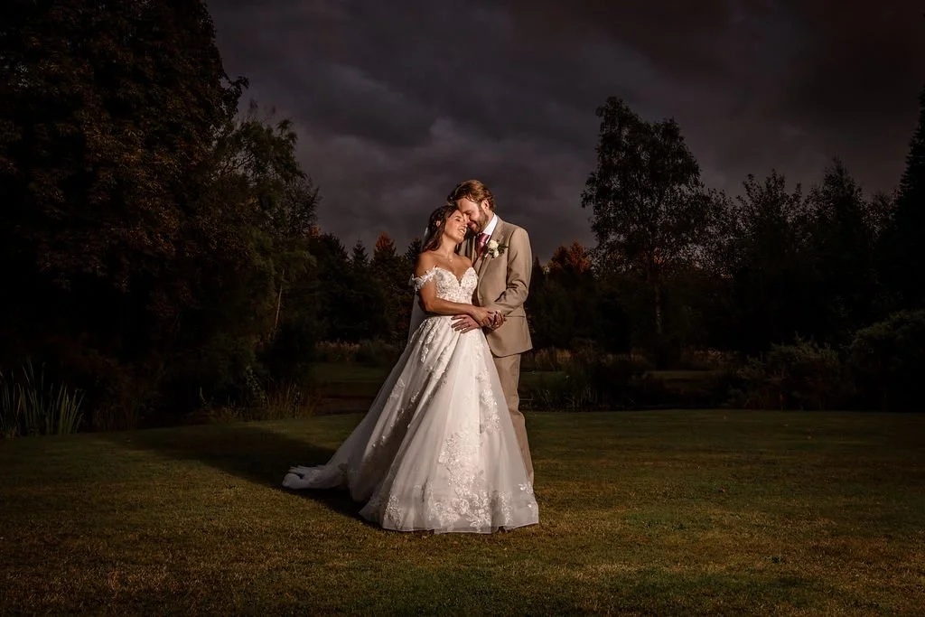 A bride and groom embracing on a lawn at night with dark, cloudy skies and trees in the background.