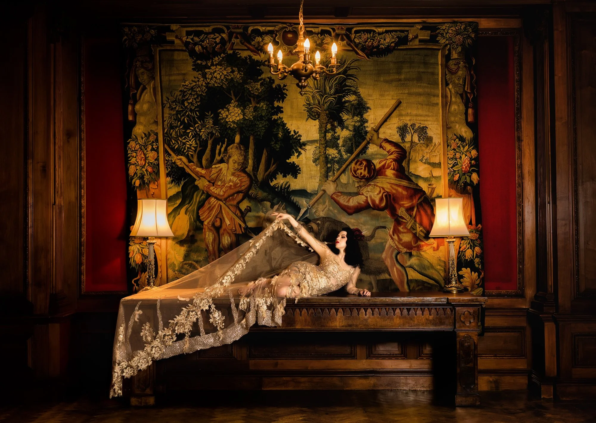 A woman in an elegant, lace wedding dress reclines on a wooden table in a dimly lit room, with a large tapestry depicting a historical scene of two men sword fighting in the background. There are two lamps with lampshades on either side of her and a chandelier hanging from the ceiling.