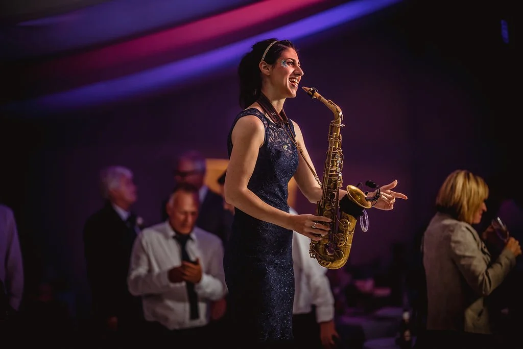 A woman in a dark blue dress playing a saxophone at a party or event, with other people in the background.