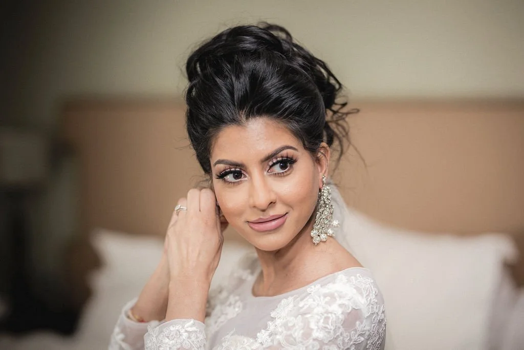 Close-up of a woman with dark hair styled in an elegant updo, wearing a white lace dress and long dangling earrings, adjusting her earring with her left hand in a softly lit room.