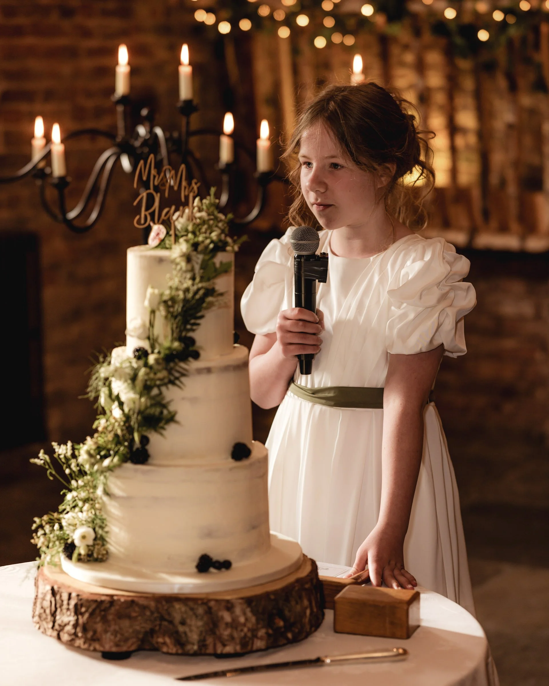 A young girl in an elegant white dress standing beside a large wedding cake, holding a microphone, at a celebration with candlelit chandeliers and warm lighting.
