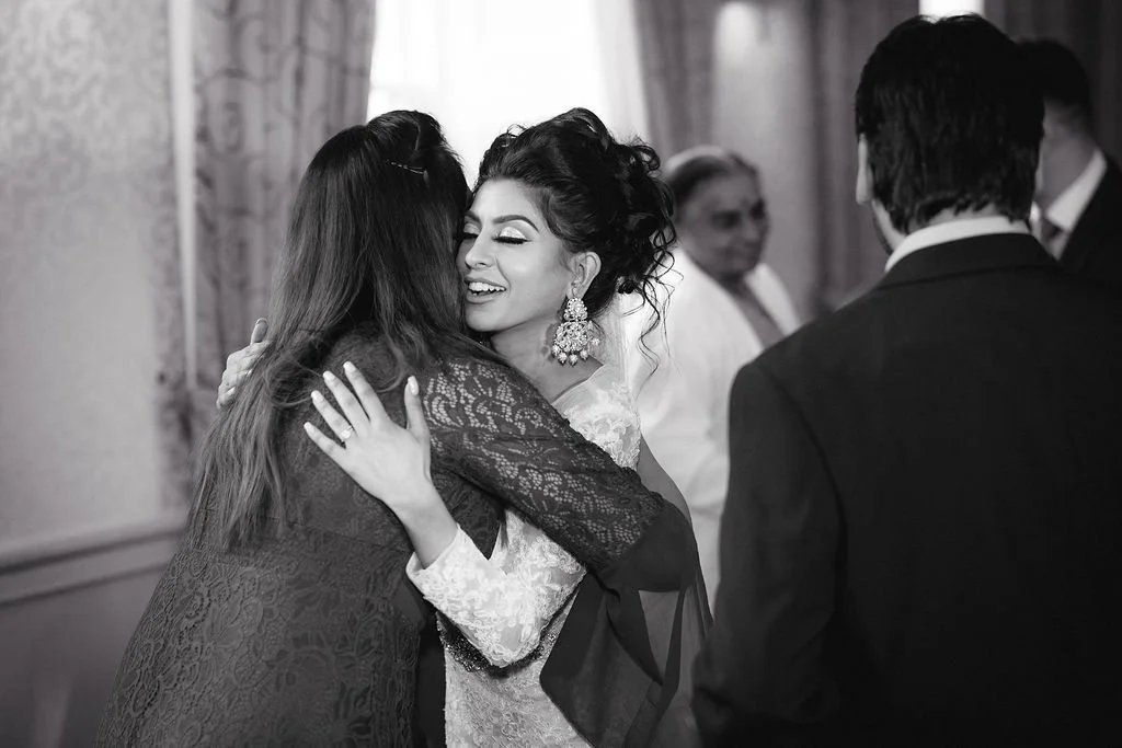 Two women hugging at a formal event, with a woman smiling and wearing earrings in the center, surrounded by men in suits and women, in a decorated indoor setting.