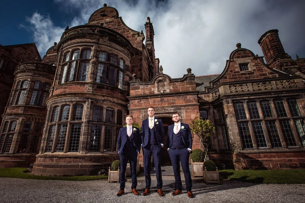 Three men in formal suits standing in front of a historic stone building with large windows, under a cloudy sky.