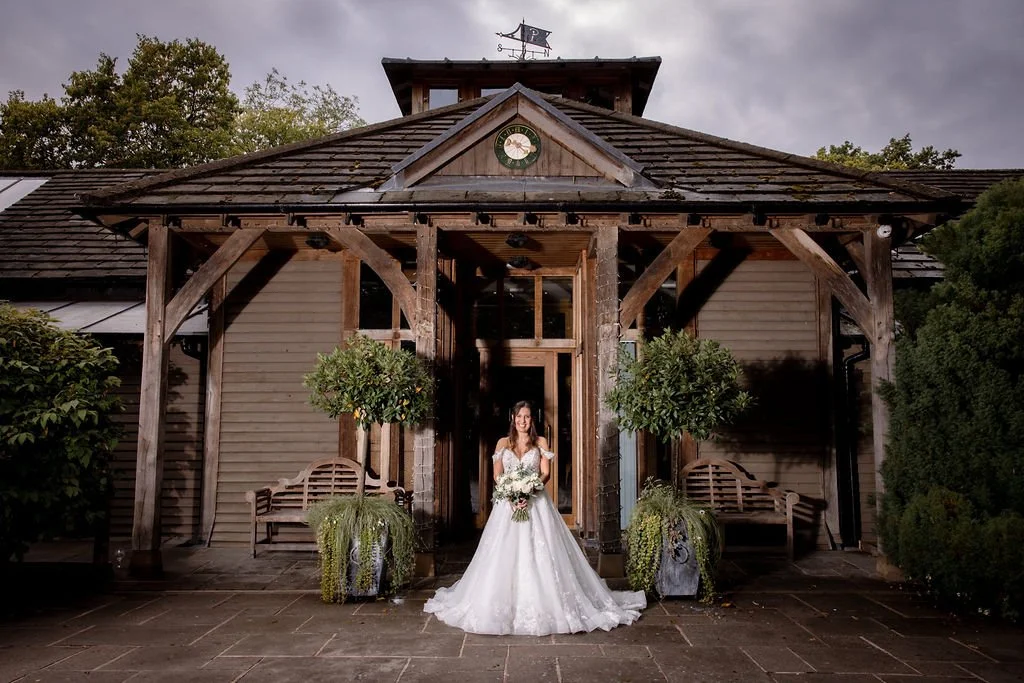 A bride in a white wedding dress holding a bouquet standing in front of a rustic wooden building with trees and cloudy sky in the background.