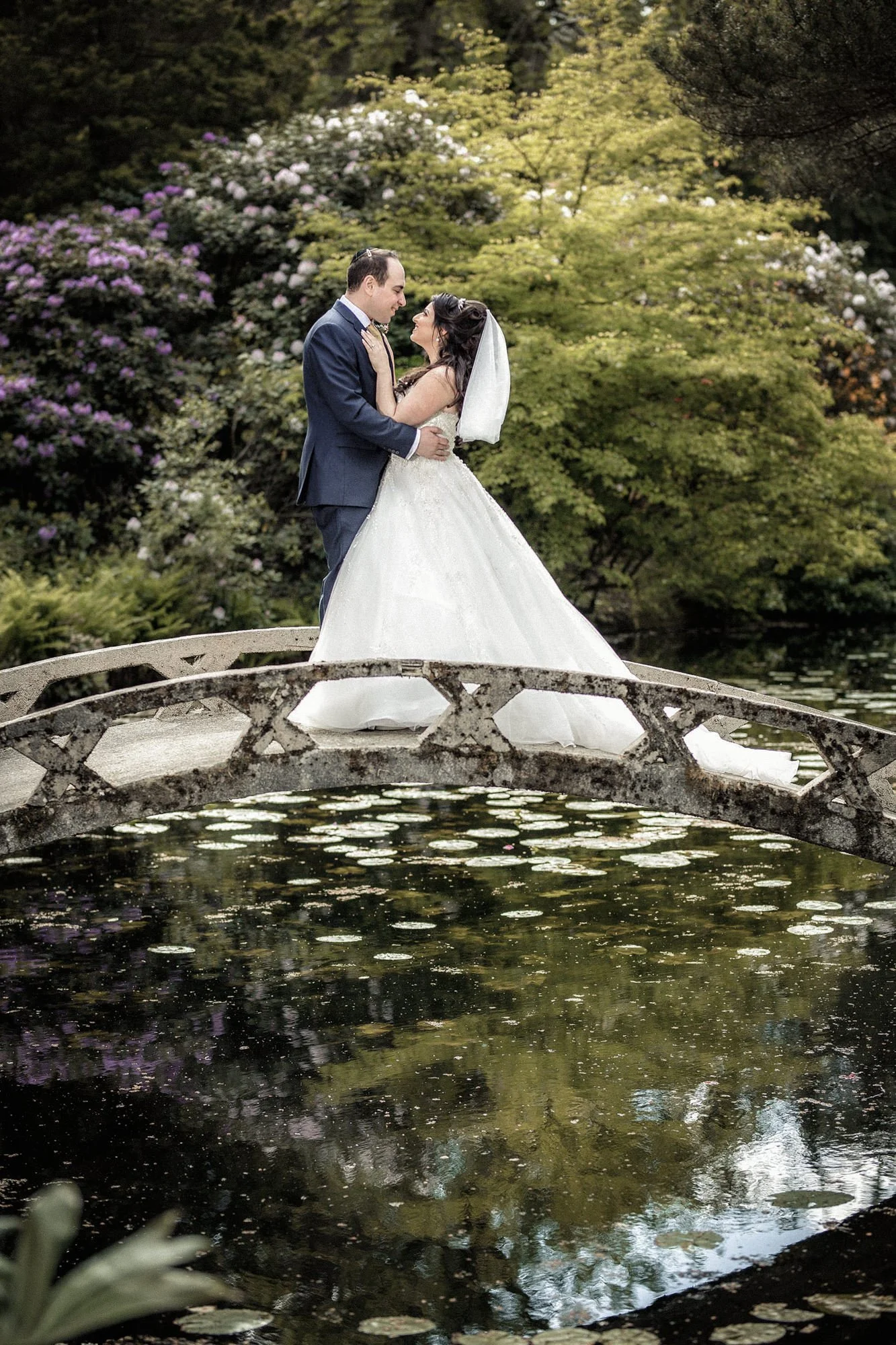 A bride and groom standing on a small stone bridge over a pond, embracing each other, surrounded by lush green trees and flowering bushes.