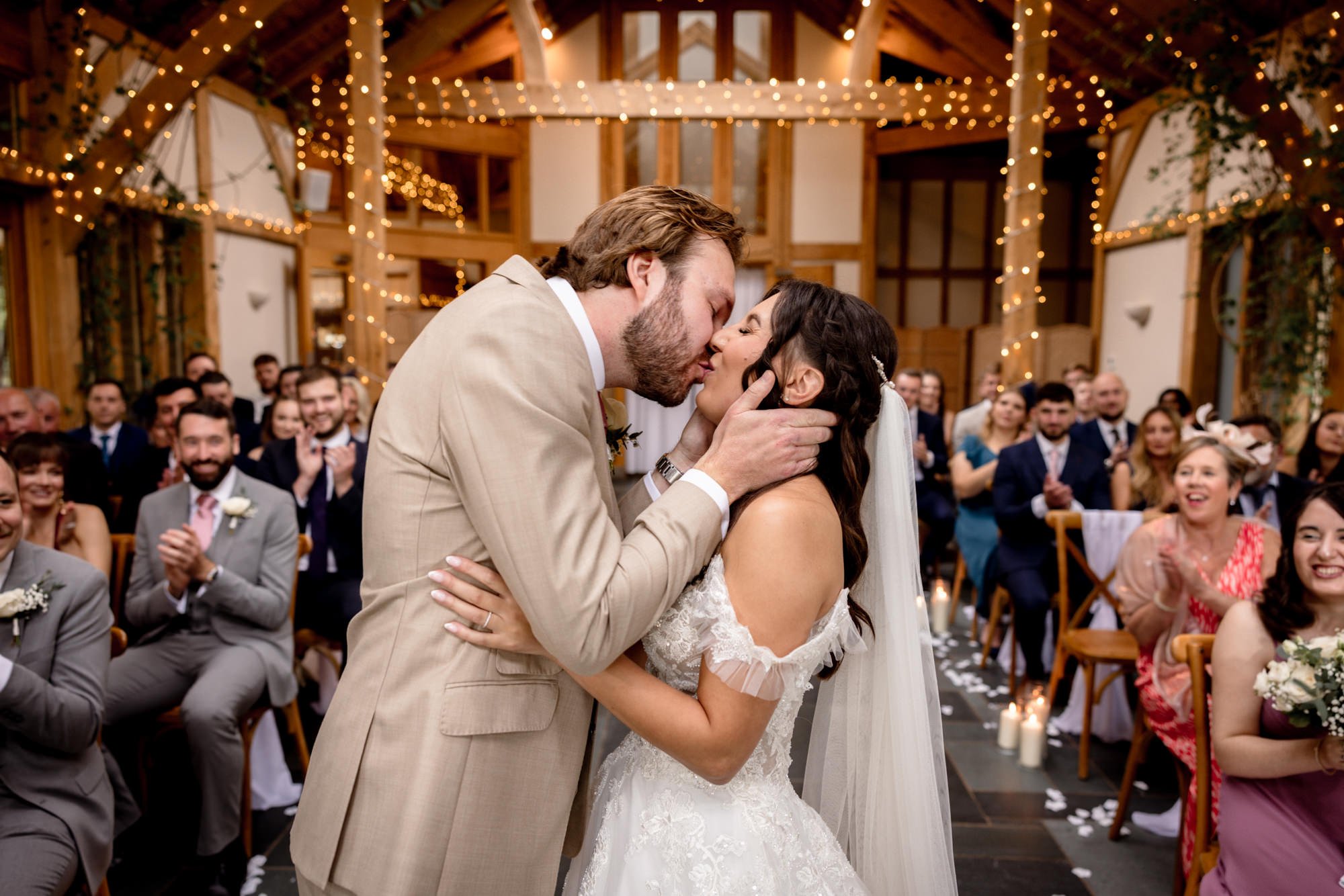 A couple shares a kiss during a wedding ceremony with guests clapping and smiling in a decorated wood-paneled venue.
